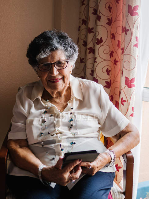 Старша жінка в сорочці і окулярах сидить на кріслі і читає електронну книгу в квартирі — стокове фото