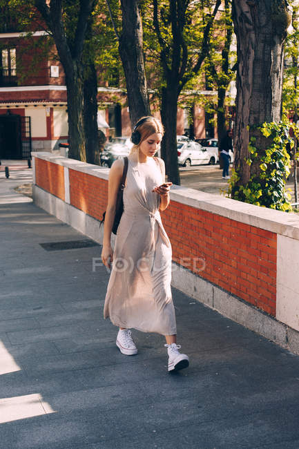 Mulher atraente em vestido de verão longo ouvir música durante a caminhada na rua à luz do dia — Fotografia de Stock