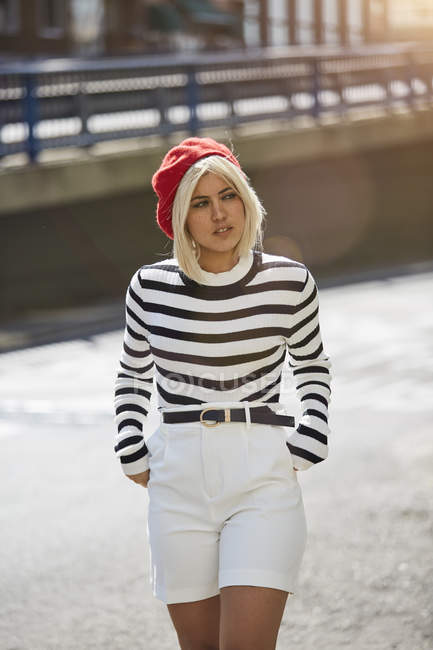 Young blonde woman in striped black and white shirt and red French cap walking on blurred city background — Stock Photo
