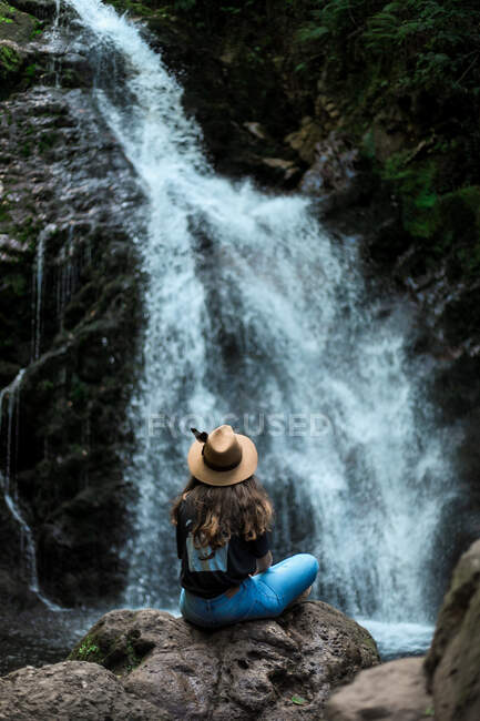 Fröhliche Reisende mit Hut lächelt und blickt in die Kamera, während sie auf nassen Felsbrocken in der Nähe des Wasserfalls sitzt — Stockfoto