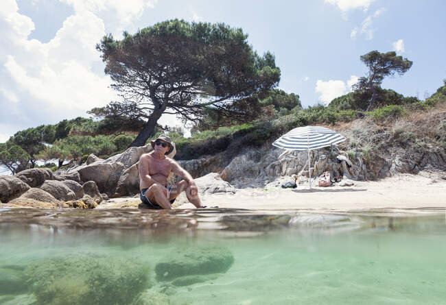 Hombre mayor relajado en sombrero acostado en la playa de arena con agua dulce descansando en la luz del sol brillante, Halkidiki, Grecia - foto de stock