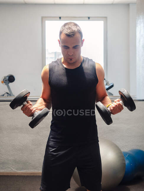 Мускулистый взрослый мужчина делает упражнения с гантелями, стоя в спортзале — стоковое фото