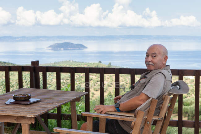 Side view of content retired man with sitting in chair at table on terrace with beautiful ocean view on background, Greece — Stock Photo