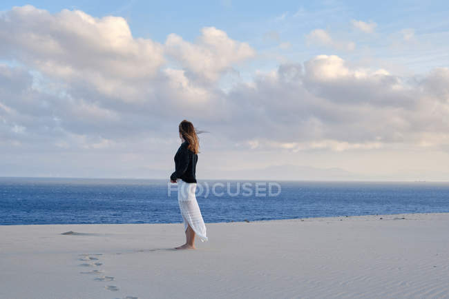 Vista lateral de la mujer de pie en el remoto desierto de arena al atardecer mirando a la cámara en Tarifa, Cádiz - foto de stock