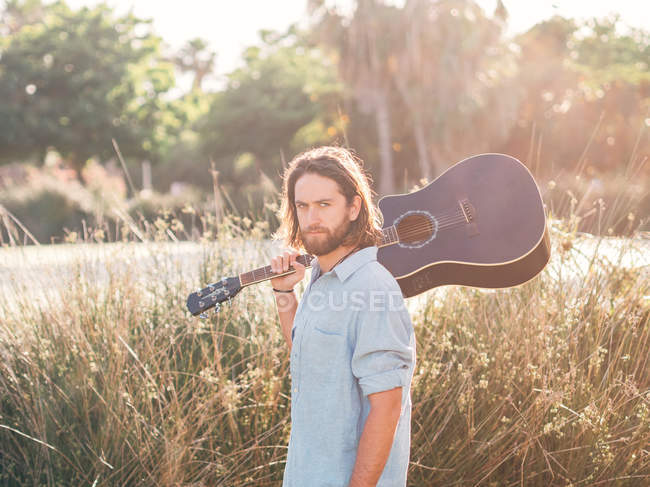 Pensive bearded hipster man walking in jungle with guitar on shoulder looking in camera — Stock Photo