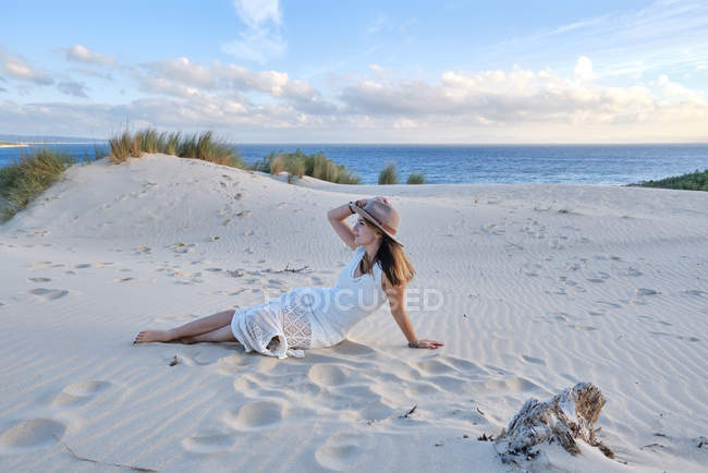Молодая красивая женщина в белом платье и шляпе лежит на песчаном пляже в Тарифе, Испания — стоковое фото
