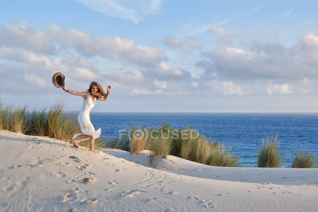 Вид сбоку женщины в белом платье с шляпой в руке, быстро бегущей по песчаному холму на пляже на фоне голубого неба в Уфе, Испания — стоковое фото
