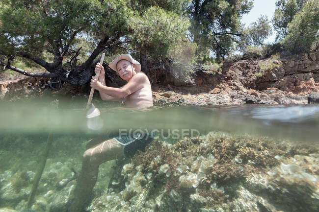 Vista lateral del anciano hombre bigote en sombrero disfrutando de agua clara estar de vacaciones y sentado bajo el agua con palo, Halkidiki, Grecia - foto de stock