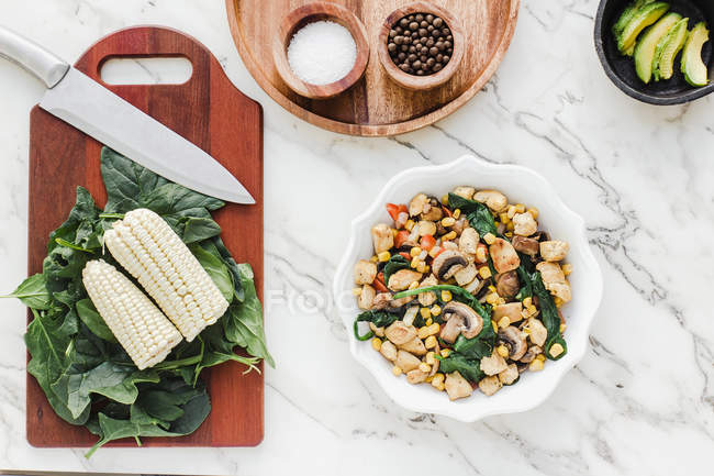 Served bowl with mushrooms, corn and cut chicken salad on table with corn leaves of greenery on cutting board and condiments — Stock Photo