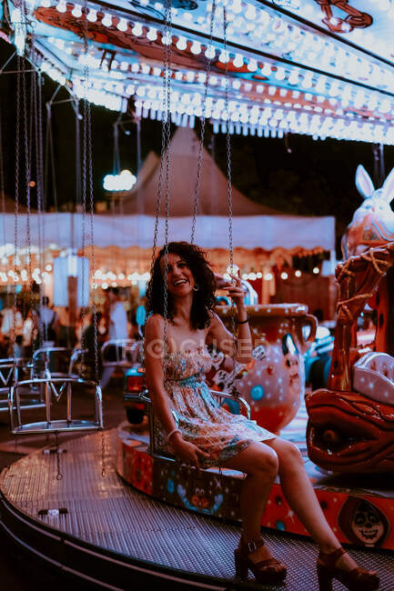 Cheerful beautiful woman enjoying ride on carousel on funfair during summer evening on blurred background — Stock Photo