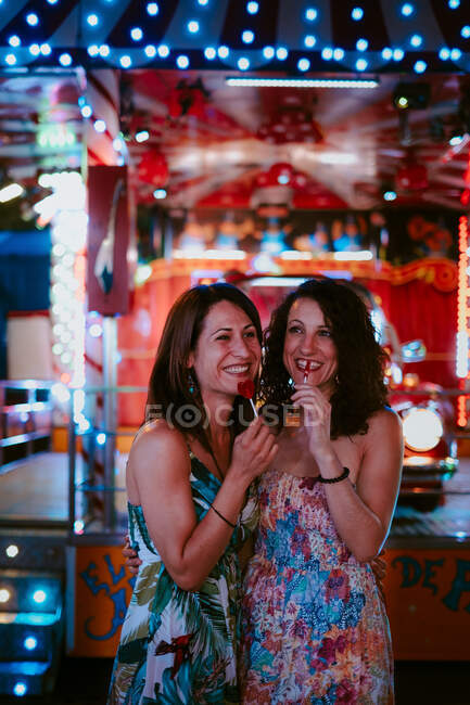 Female friends with lollipops bonding in amusement park on warm summer evening looking away on blurred background — Stock Photo