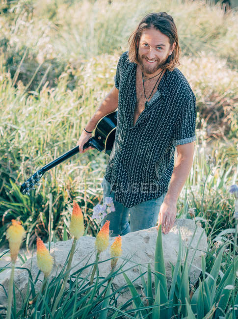 Homme hipster barbu souriant marchant dans la jungle avec la guitare regardant à la caméra — Photo de stock