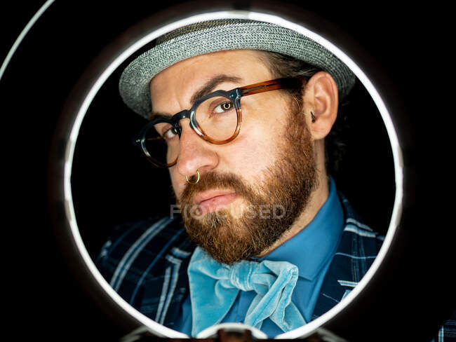 Bel homme barbu dans des lunettes et regardant la caméra sur fond noir et la lumière du cercle — Photo de stock