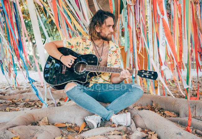 Bearded man sitting under decorated tree playing guitar — Stock Photo