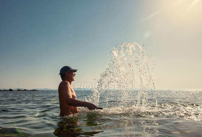 Vista lateral del hombre curtido maduro en gorra disfrutando del agua mientras salpica en el día soleado brillante en Halkidiki, Grecia - foto de stock