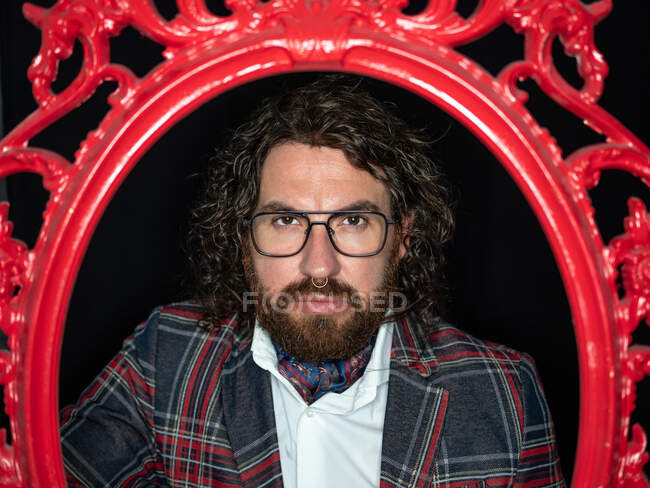 Serious impresionante hombre bien vestido con el pelo rizado y la barba posando en marco estampado rojo sobre fondo negro - foto de stock