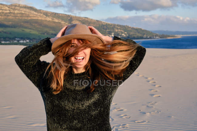 Mujer viajera alegre en sombrero de pie en el desierto de arena remota en la puesta de sol mirando a la cámara en Tarifa, Cádiz - foto de stock