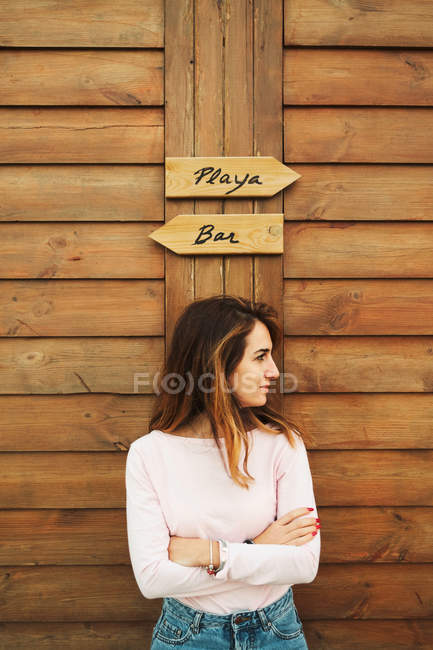 Fiduciosa bella donna con le mani incrociate appoggiata su una parete di legno con due segni che distolgono lo sguardo a Tarifa, Spagna — Foto stock