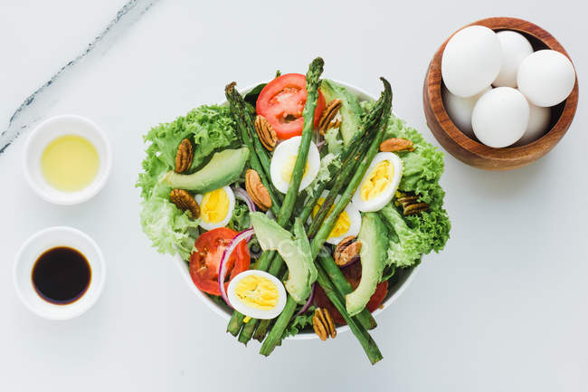 Served bowl of salad with asparagus, eggs, avocado, tomatoes, walnuts and greenery on table with condiments, sauces and eggs — Stock Photo