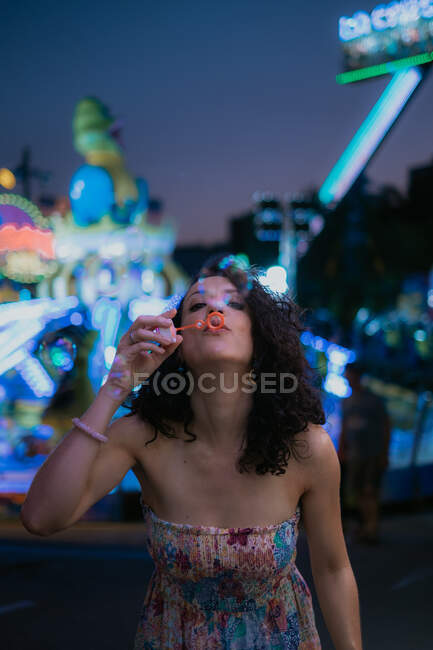 Mulher em vestido de verão elegante desfrutando de tempo à noite no parque de diversões e soprando bolhas no fundo borrado — Fotografia de Stock