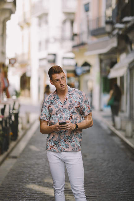Bonito macho em camisa havaiana de pé na rua, usando telefone celular e olhando para longe — Fotografia de Stock