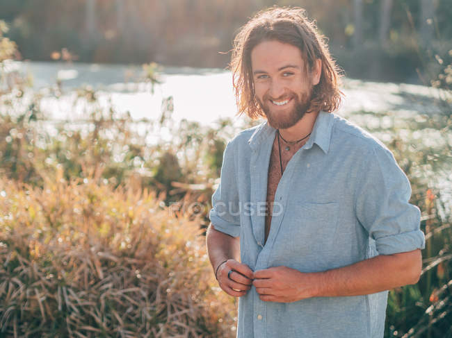 Jeune homme hipster barbu joyeux déboutonnant chemise bleue se préparant à nager dans la rivière — Photo de stock