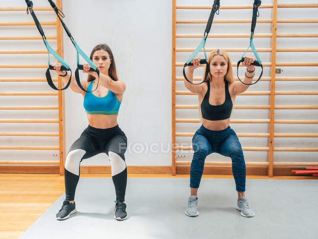Junge selbstbewusste Frauen machen Kniebeugen beim Hängetraining mit Seilen in einem modernen Fitnessstudio und schauen in die Kamera — Stockfoto