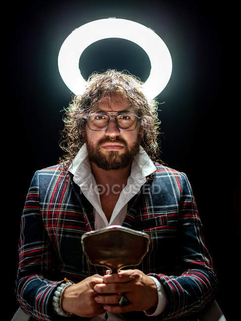 Stylish well dressed man with beard holding beauty mirror and looking at camera on black background under glowing bright fake nimbus — Stock Photo