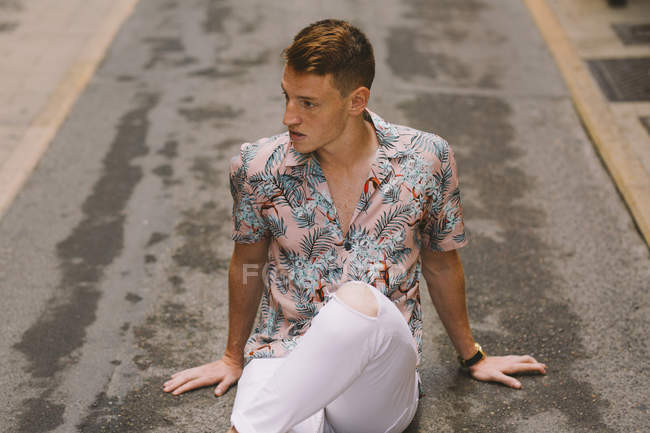Hombre guapo en camisa hawaiana sentado en asfalto en la calle con las piernas cruzadas y mirando hacia otro lado - foto de stock
