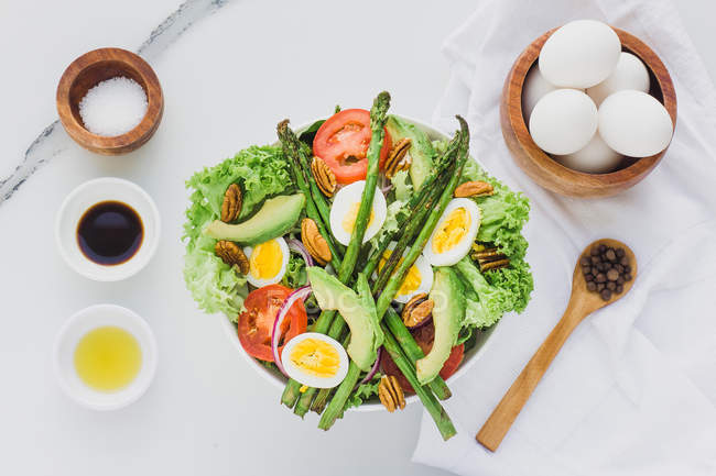 Served bowl of salad with asparagus, eggs, avocado, tomatoes, walnuts and greenery on table with condiments, sauces and eggs — Stock Photo