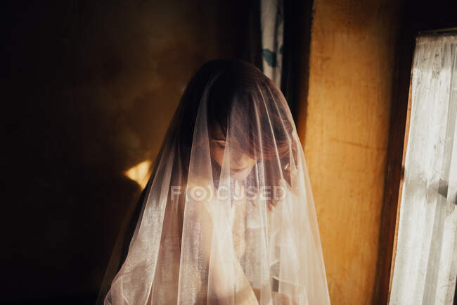 Голая чувственная женщина, покрытая прозрачной белой тканью, сидит у окна под солнечным светом — стоковое фото