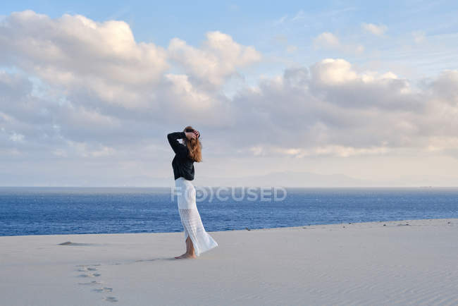 Vista lateral de la mujer de pie en el remoto desierto de arena al atardecer mirando a la cámara en Tarifa, Cádiz - foto de stock