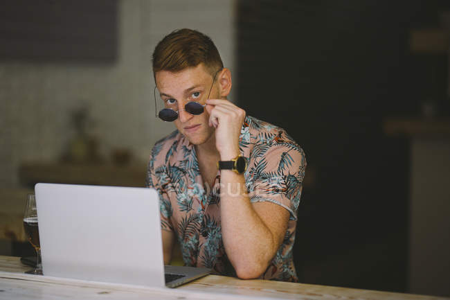 Focused young man working on laptop at table in coffee shop and looking through sunglasses in camera — Stock Photo