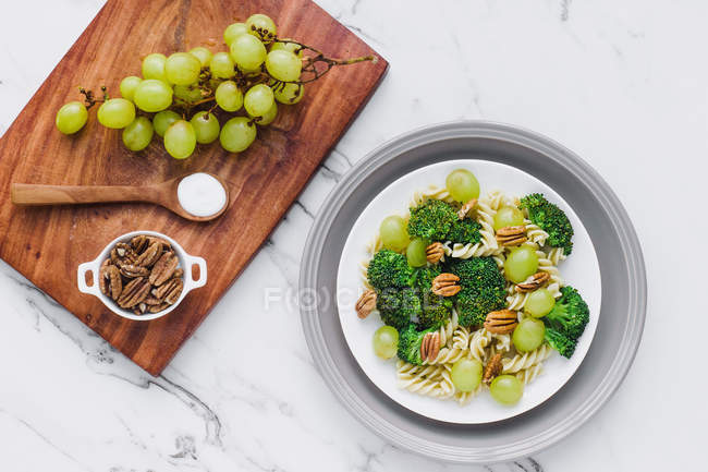 Servido tigela de salada com macarrão, brócolis e nozes na mesa com uvas e sal na tábua de corte — Fotografia de Stock