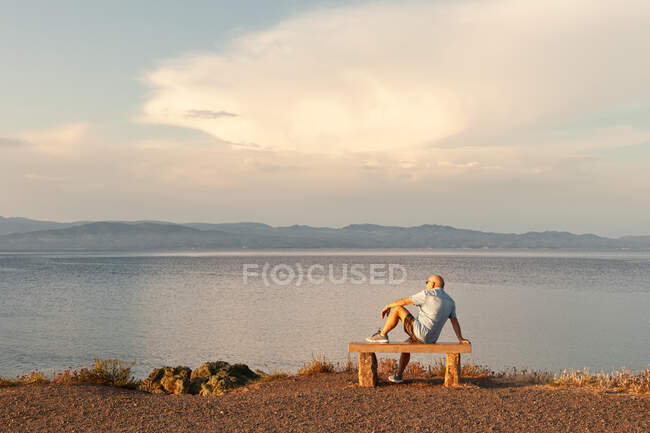Vista trasera del anciano sentado en el banco y disfrutando de un paisaje marino tranquilo en el tiempo de la puesta del sol, Halkidiki, Grecia - foto de stock