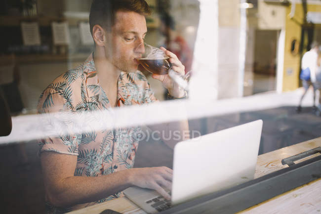 Focalizzato giovane uomo utilizzando il computer portatile a tavola durante le finestre e bere birra nel caffè — Foto stock