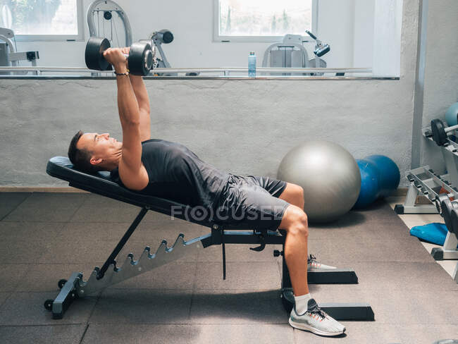Vue latérale d'un homme adulte musclé faisant de l'exercice avec des haltères tout en étant allongé sur un banc dans une salle de gym — Photo de stock
