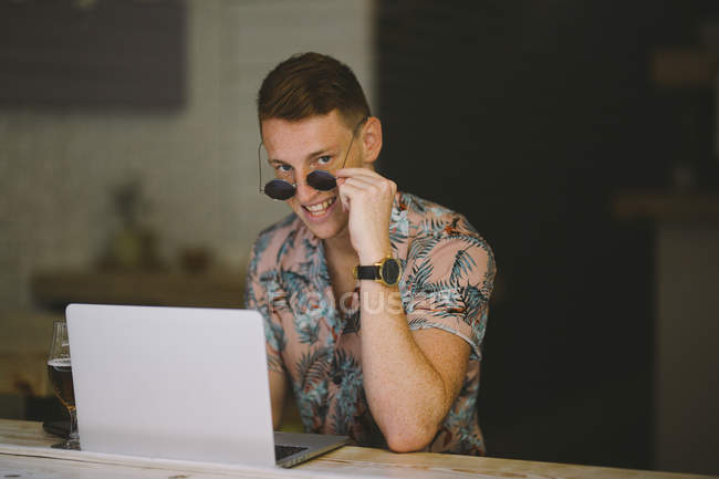 Jovem focado trabalhando no laptop à mesa no café, olhando através de óculos de sol na câmera e sorrindo — Fotografia de Stock