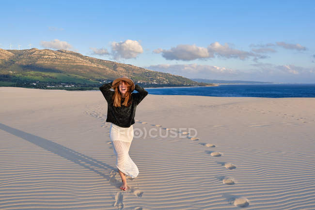 Fröhliche reisende Frau mit Hut steht in einer abgelegenen Sandwüste bei Sonnenuntergang und blickt in die Kamera in Tarifa, Spanien — Stockfoto