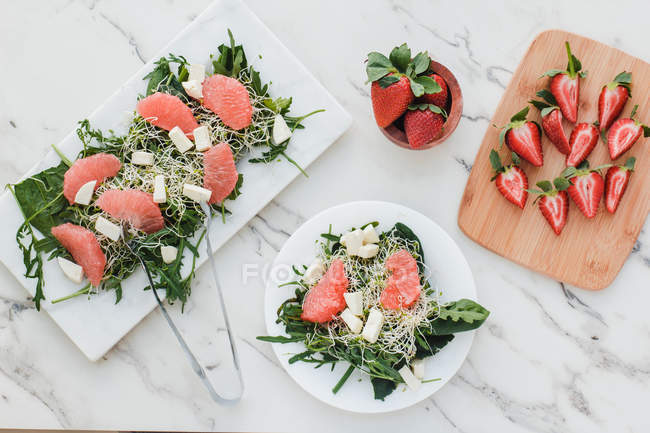 Vue du dessus des bols avec des fraises, du pamplemousse et de la salade de fusée sur la table servie sur des planches — Photo de stock
