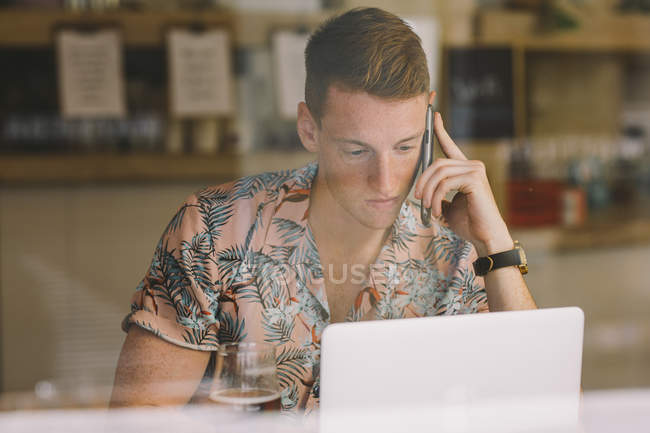 Орієнтований молодий чоловік розмовляє по смартфону, використовуючи ноутбук за столом по всіх вікнах у кав'ярні — стокове фото