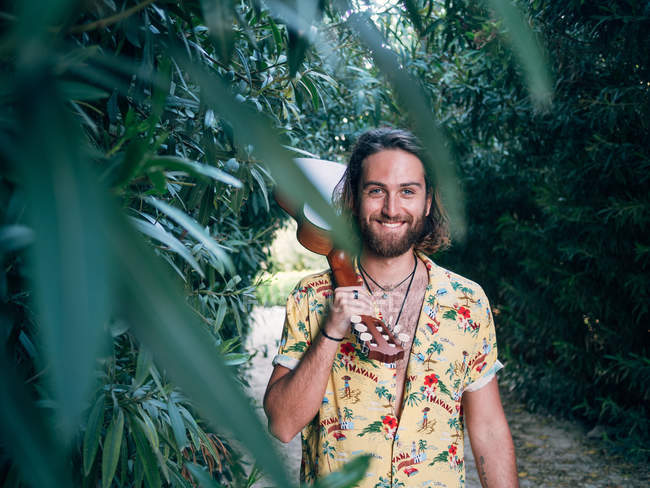 Pensive bearded hipster man walking in jungle with ukulele looking in camera — Stock Photo