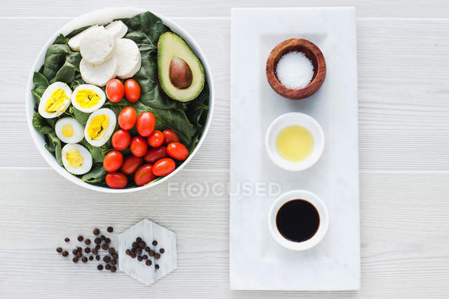 Served bowl of salad with spinach, eggs, avocado, tomatoes and mozzarella cheese on table with condiments and sauces — Stock Photo