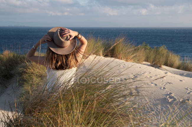 Vista posterior de la mujer con sombrero sentado en la colina de arena mirando pintorescas vistas al mar en Tarifa, Cádiz - foto de stock