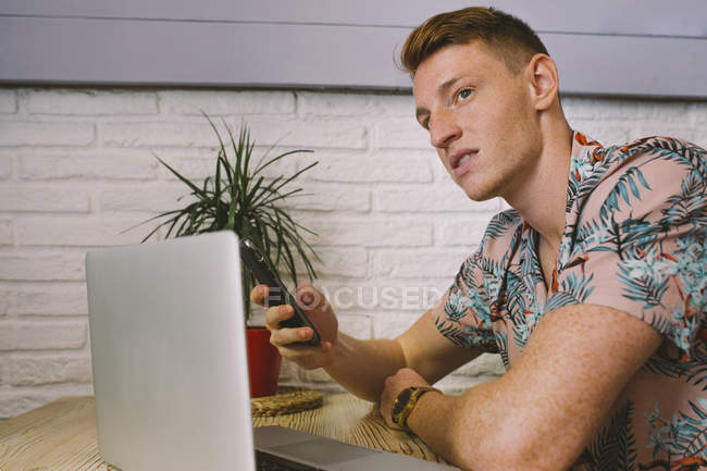 Uomo concentrato utilizzando smartphone mentre si utilizza il computer portatile a tavola in caffetteria — Foto stock