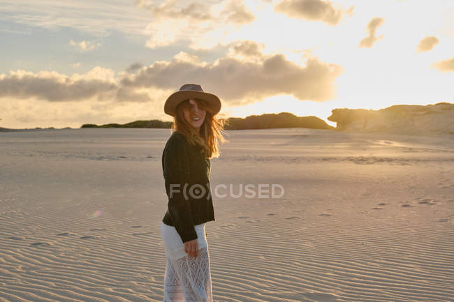 Веселий подорожній жінка у капелюсі стоїть у піщаній пустелі на заході сонця і дивиться на камеру в Таріфі (Іспанія). — стокове фото