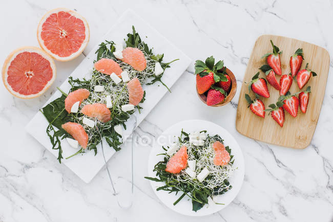 Vue du dessus des bols avec des fraises, du pamplemousse et de la salade de fusée sur la table servie sur des planches — Photo de stock