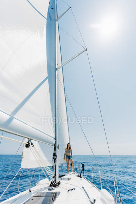 Vue latérale de la femme voyageuse portant un maillot de bain debout sur le côté du yacht en mer regardant la caméra — Photo de stock