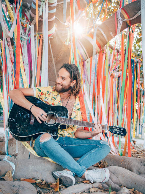 Бородатый мужчина сидит под украшенным деревом и играет на гитаре — стоковое фото