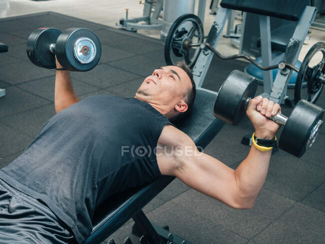 Visão lateral do homem adulto muscular fazendo exercício com halteres enquanto deitado no banco no ginásio — Fotografia de Stock